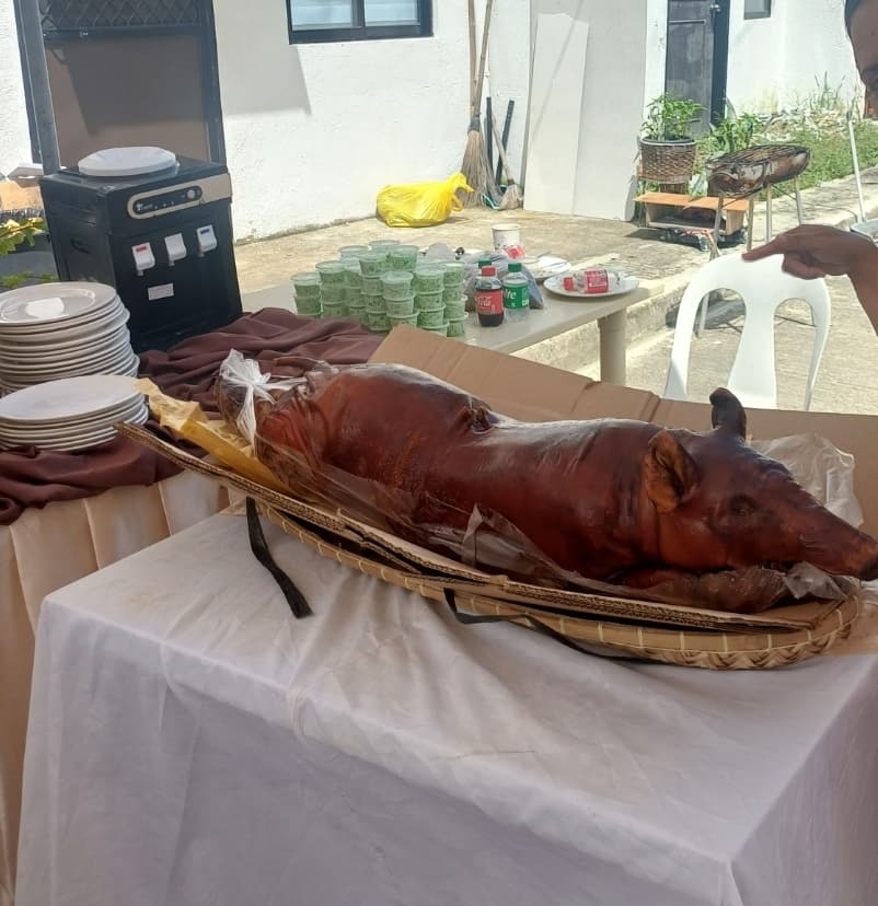 A whole roast lechon, uncarved, resting in a woven basket on a draped table at an outdoor gathering