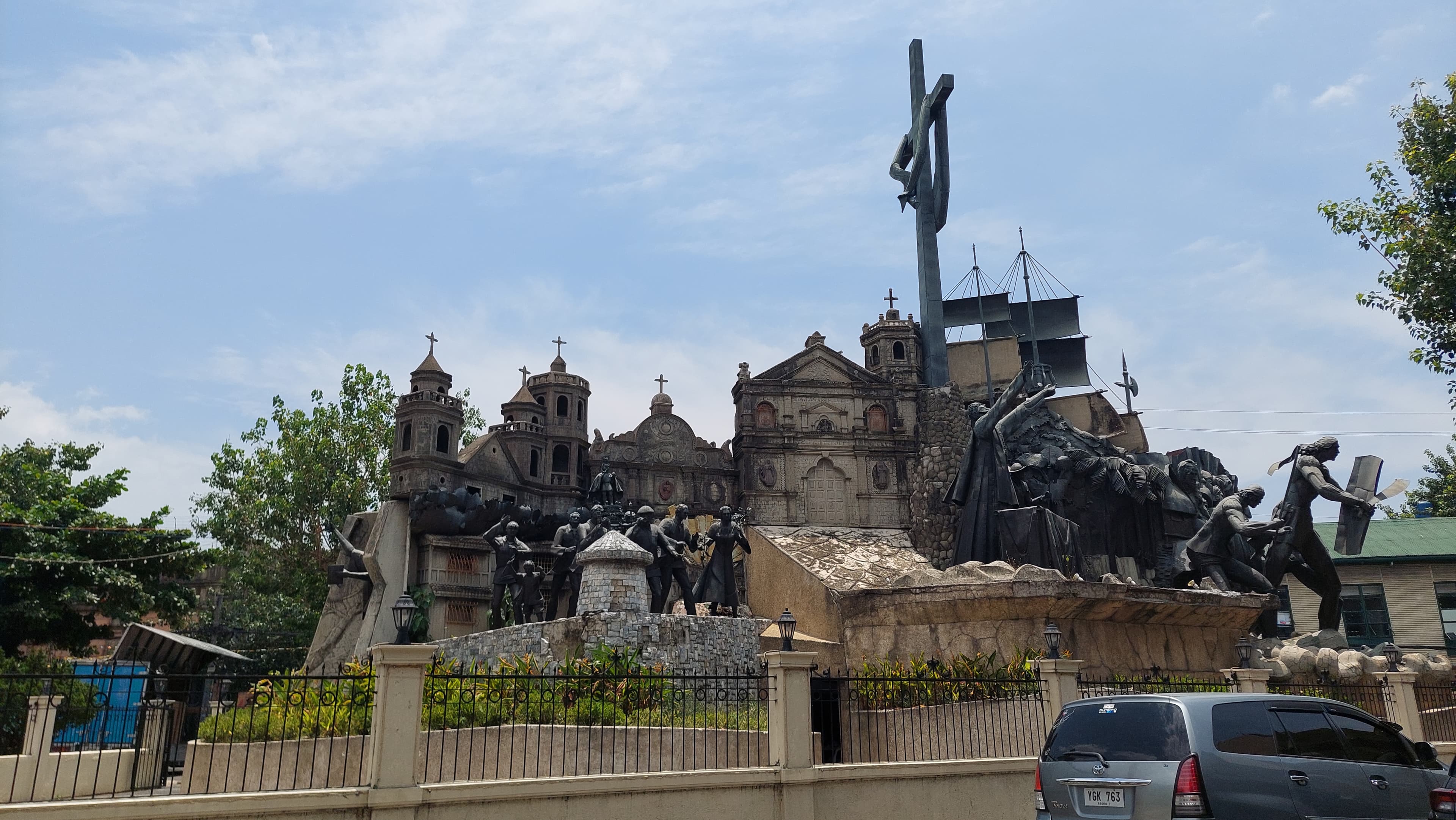 The monument's crowd of dark bronze figures massed around a stone church facade, a galleon's masts and a towering cross, behind the low fence that rings it