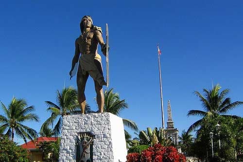 The bronze statue of Lapu-Lapu raised on a pale stone plinth, palms and the Magellan marker behind it