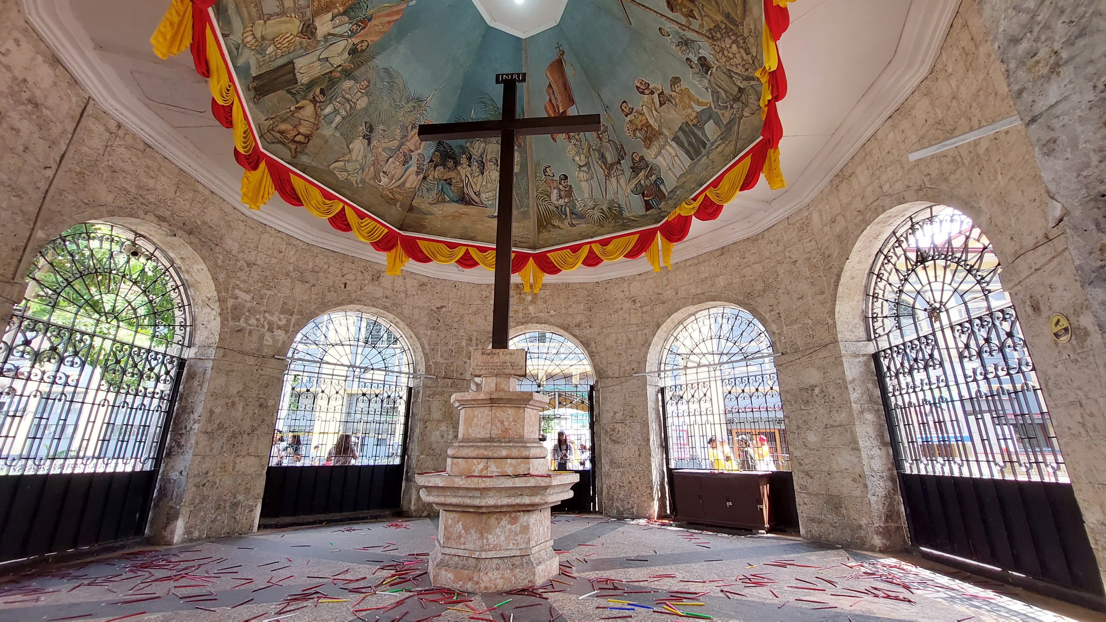 The dark wooden cross standing beneath the painted ceiling of its kiosk, the mural above it showing the 1521 planting
