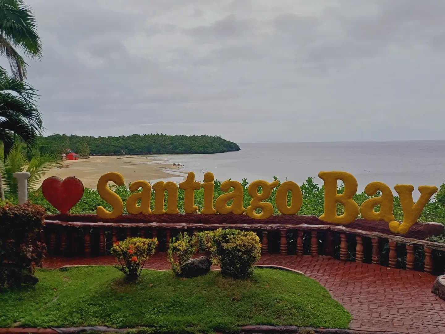 A painted "I love Santiago Bay" sign on a clifftop terrace, with the bay's pale beach and wooded headland behind it