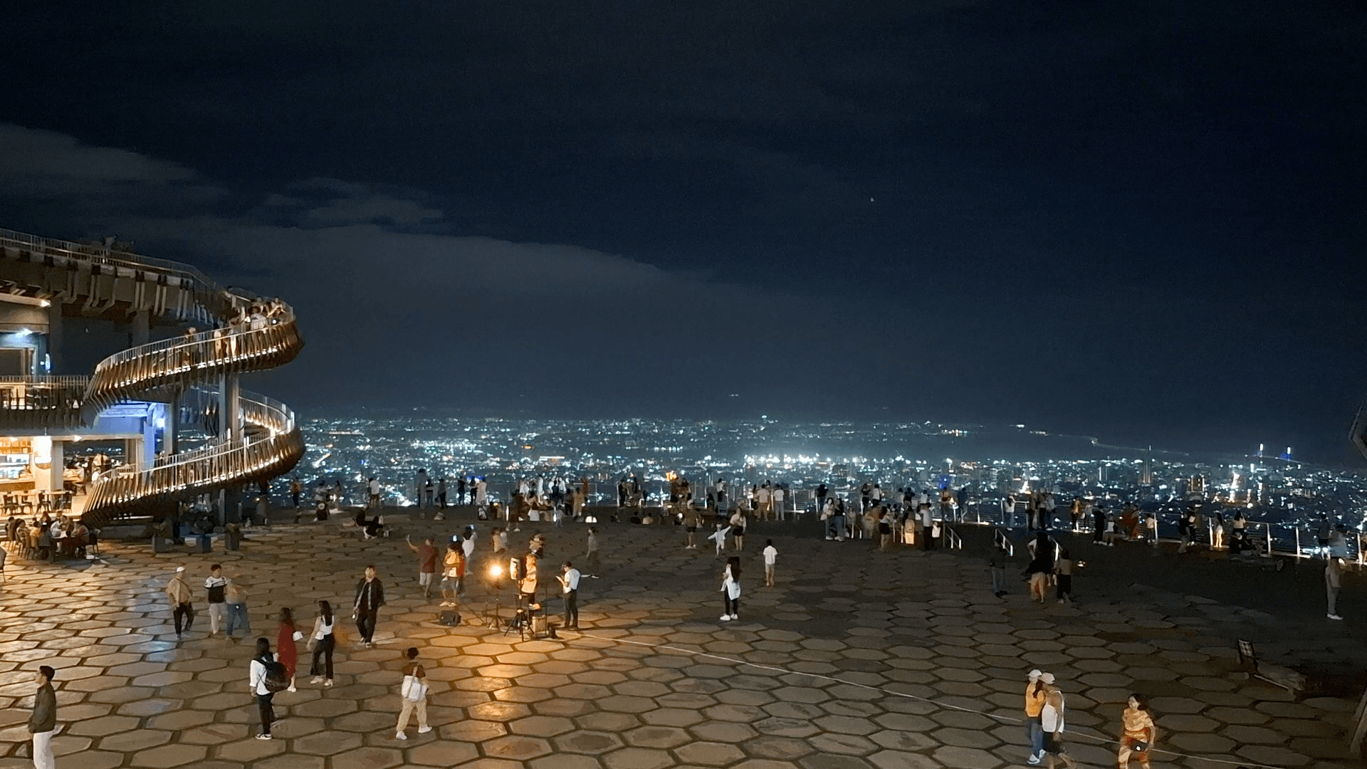 Visitors spread across the paved viewing deck after dark, the lights of Metro Cebu below and a lit spiral staircase at the left