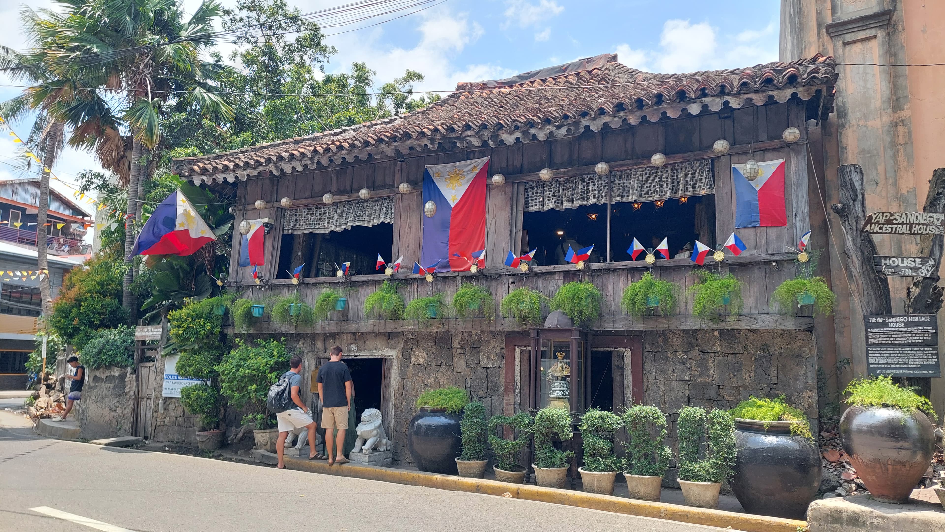 The house's dark timber upper storey and tiled roof over coral-stone walls, Philippine flags strung along the balcony and clay pots lining the street below